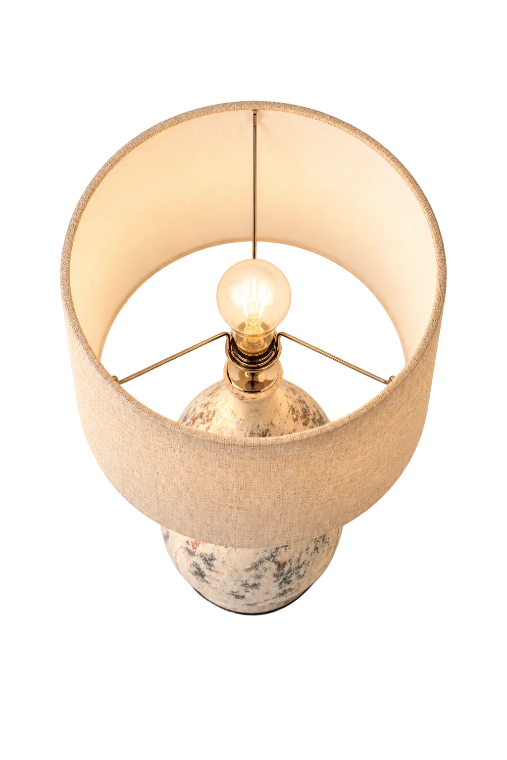 A table lamp with a warm beige fabric drum shade, a patterned ceramic base, and a glowing light bulb, photographed against a neutral light gray background.